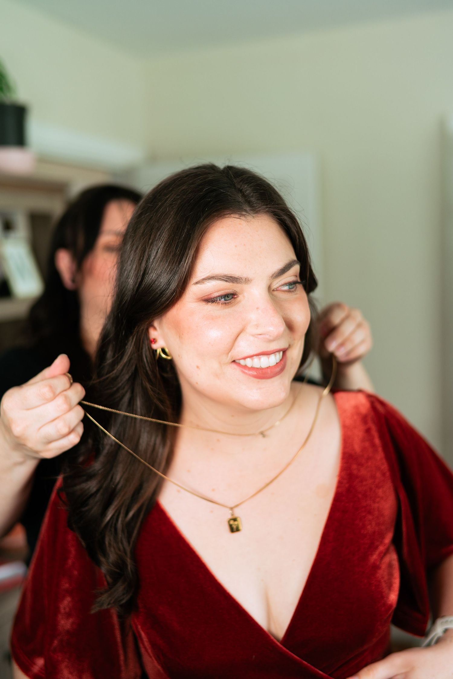 A person in a luxurious red velvet dress smiles warmly while someone helps style their long dark hair.