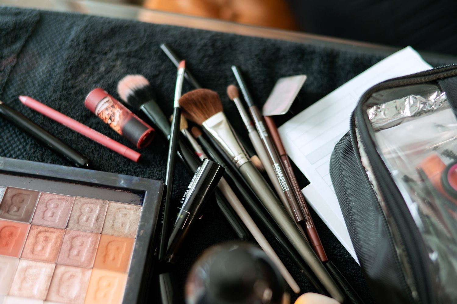 A collection of makeup brushes and cosmetic products scattered on a black surface with an eyeshadow palette visible.