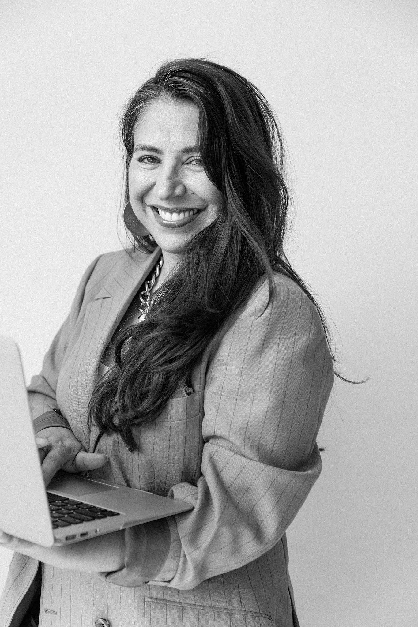 Professional in a pinstripe blazer holds laptop while smiling against white background in black and white portrait.