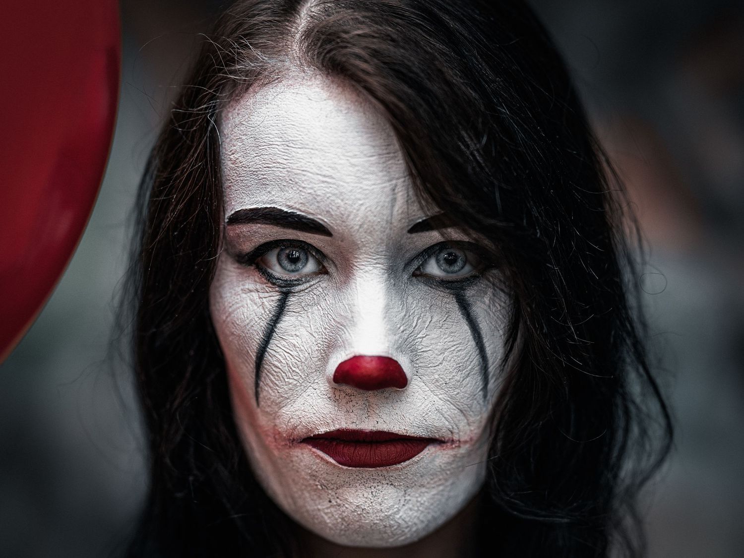 Haunting clown makeup featuring black tear streaks, a red nose, dark hair and red lipstick in dramatic lighting.