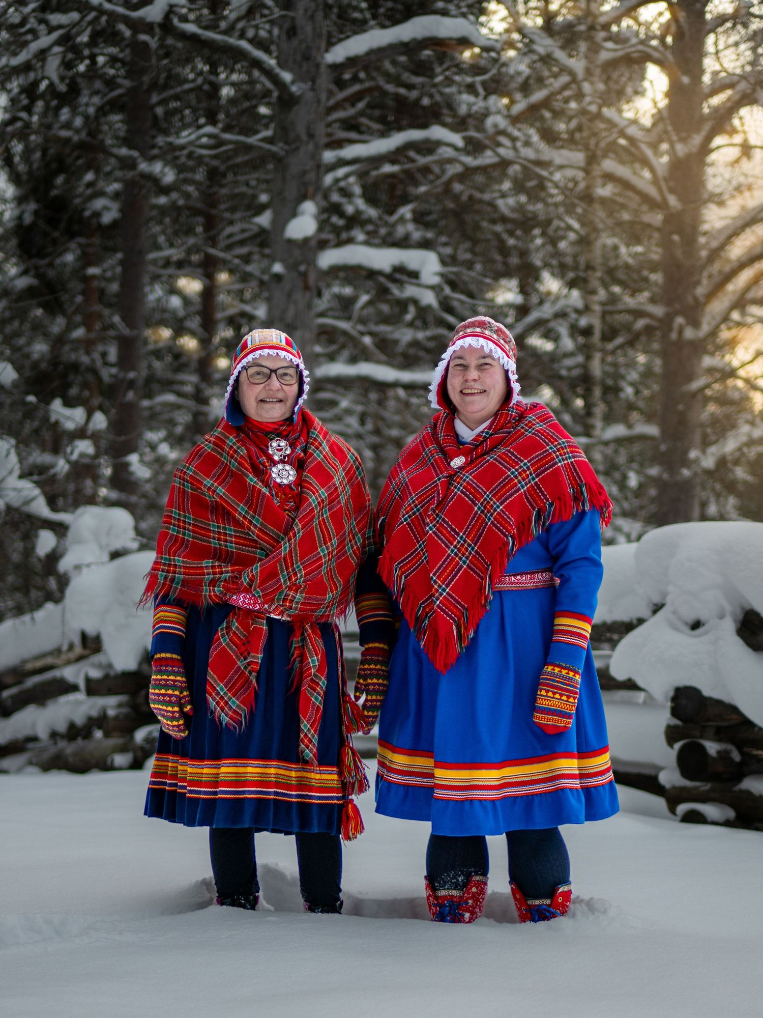 Two people wearing traditional Sami clothing pose in a snowy winter forest landscape.