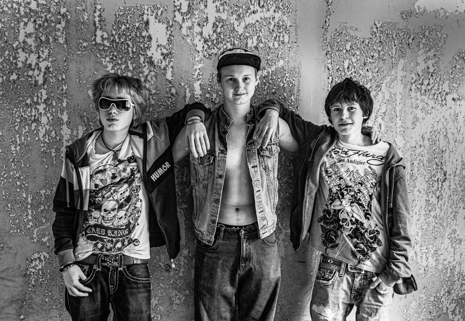 Tre punkmusiker poserar mot en grusig vägg iklädda trasiga kläder och svartvita jeans.
