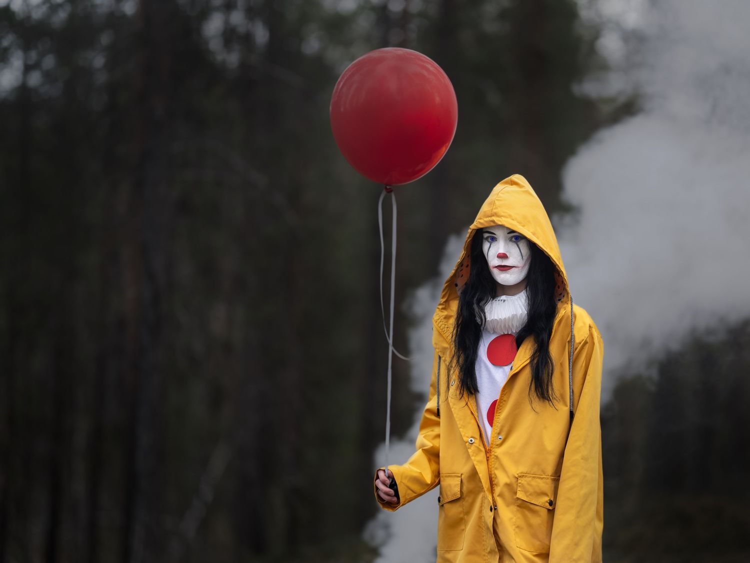 Spöklik clownkaraktär i gul regnrock som håller röd ballong mot dimmig skogsbakgrund.