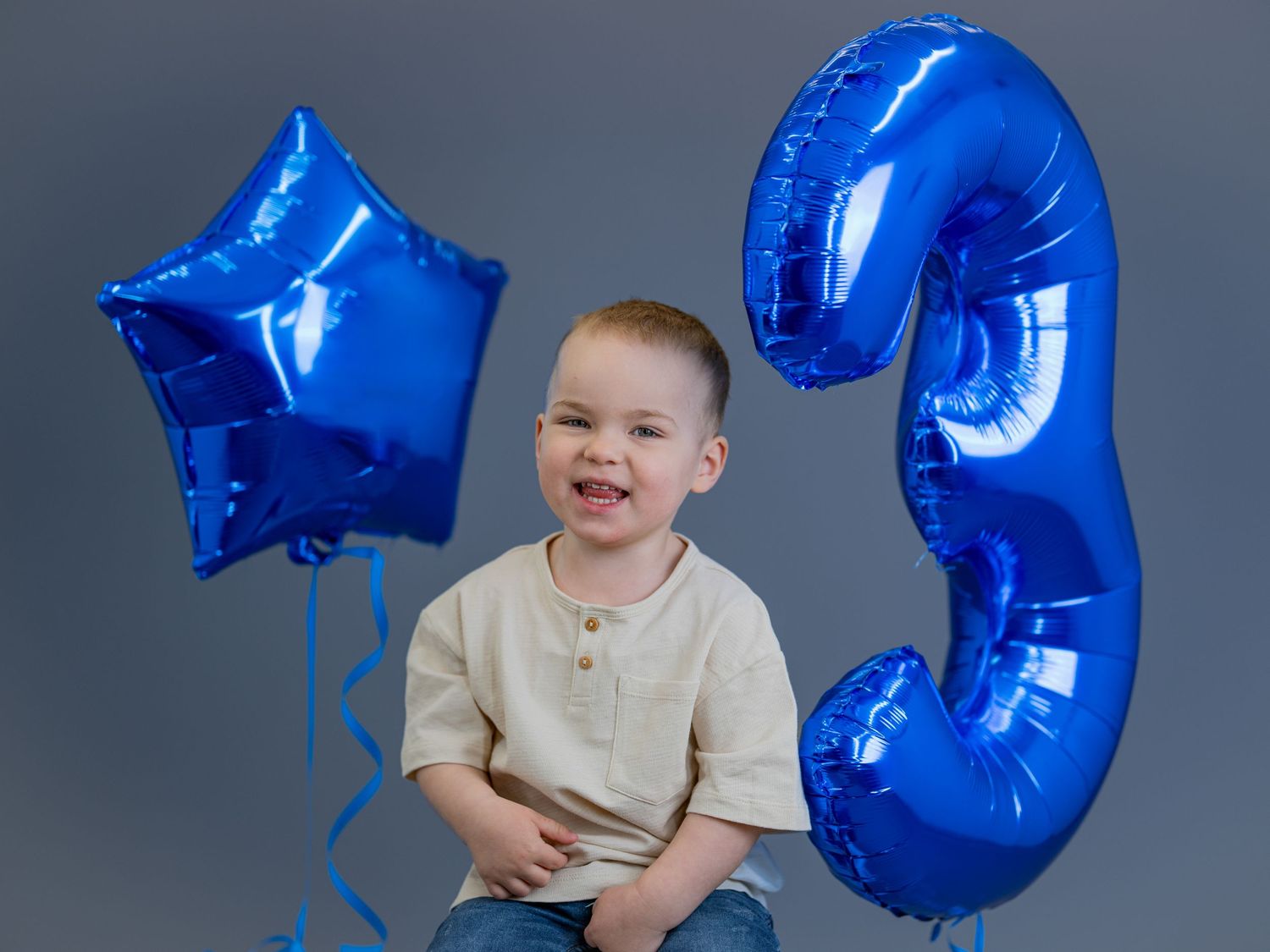 A young child sits with bright blue star and number three balloons against a grey studio backdrop.