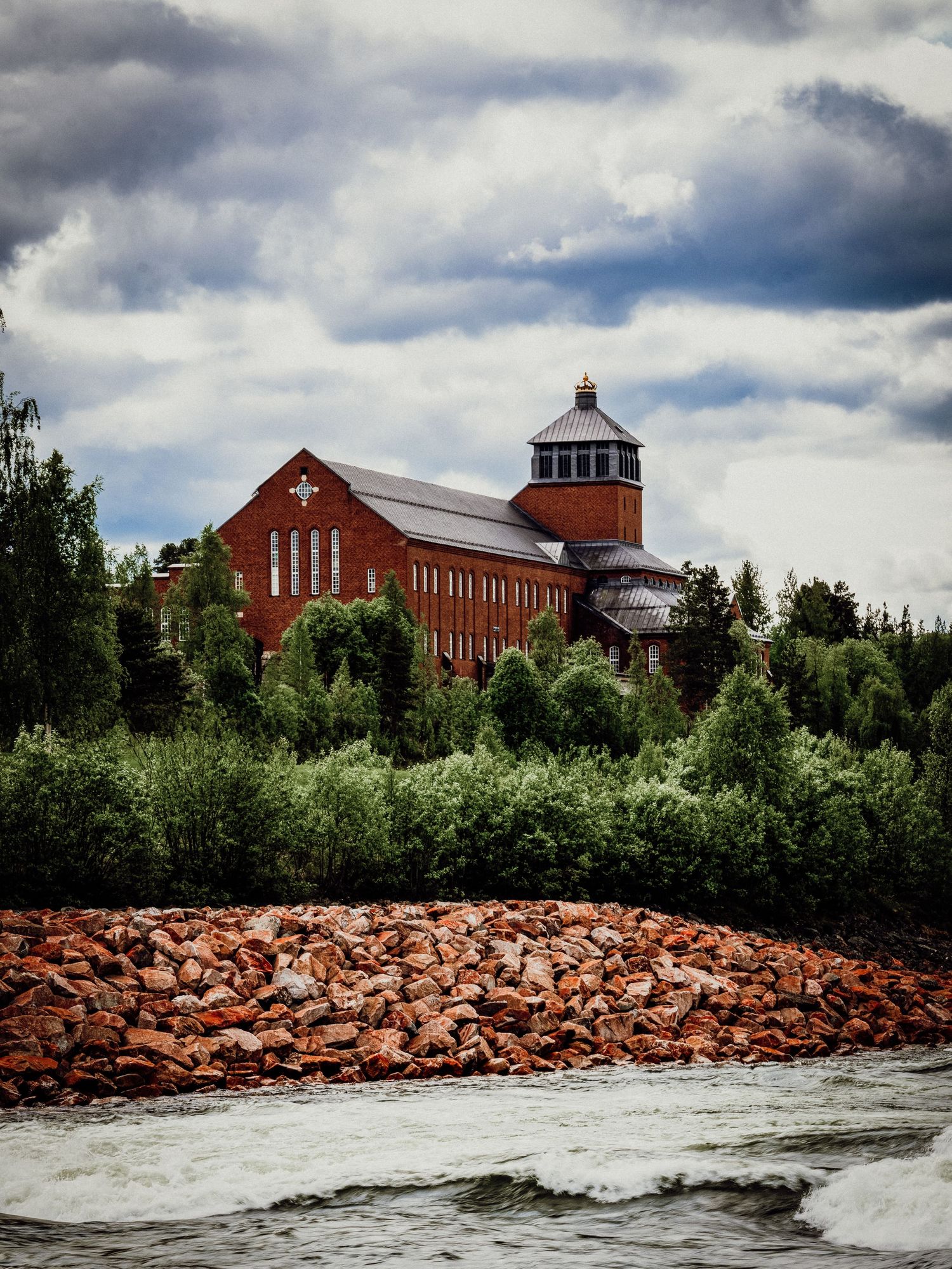 Red brick church building sits above rocky shoreline along rushing river under dramatic cloudy sky.