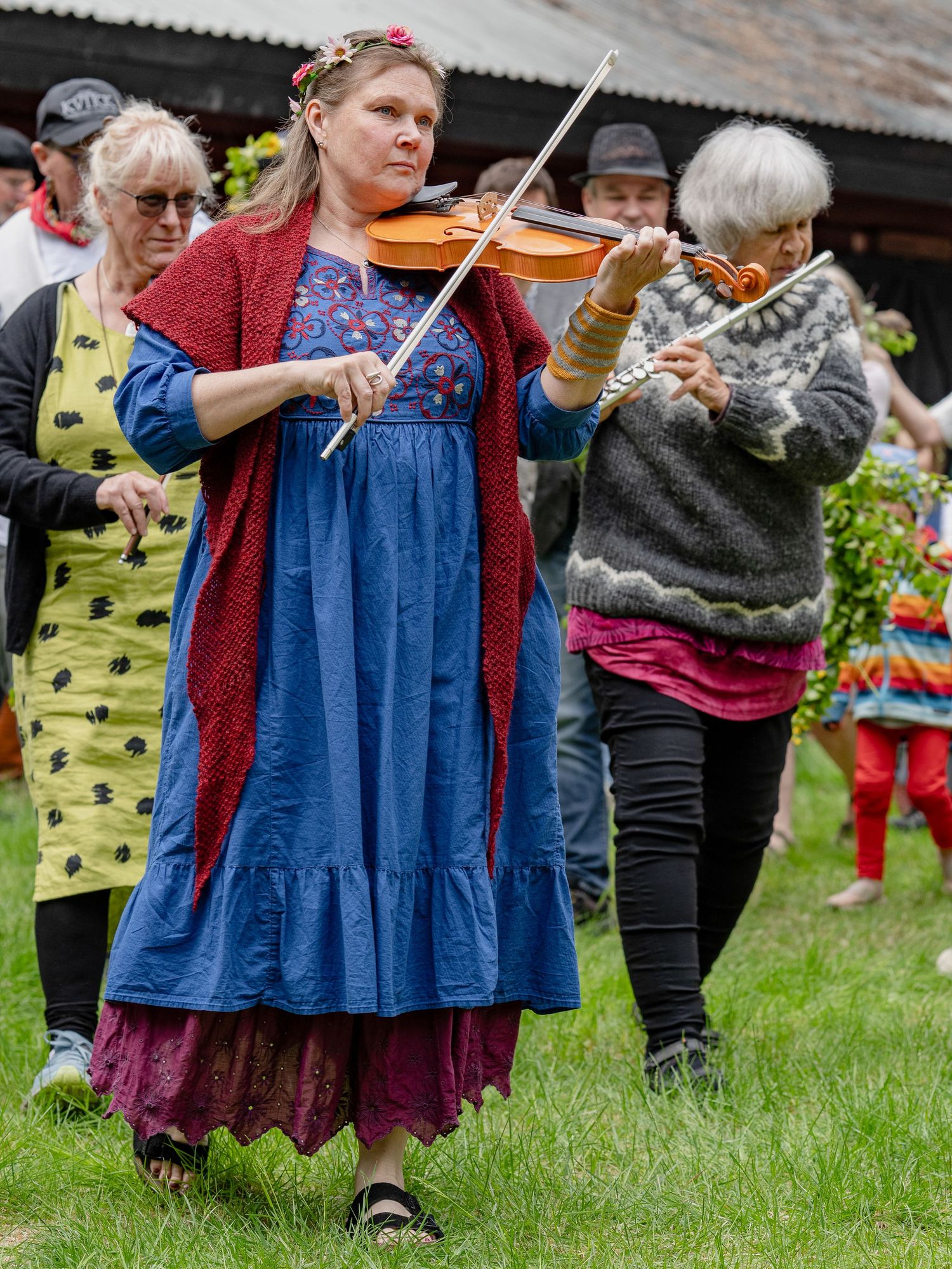 Musicians perform traditional folk music outdoors with violin and flute during a festive gathering on grass.