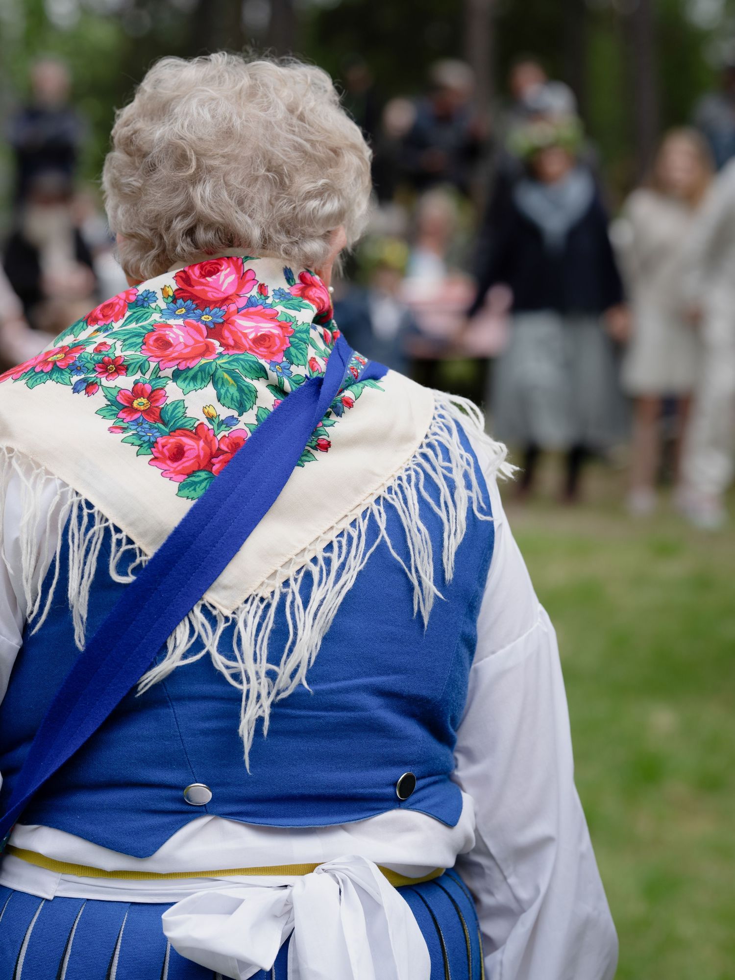 A person wearing traditional Swedish folk costume with floral shawl and blue vest seen from behind at an outdoor gathering.