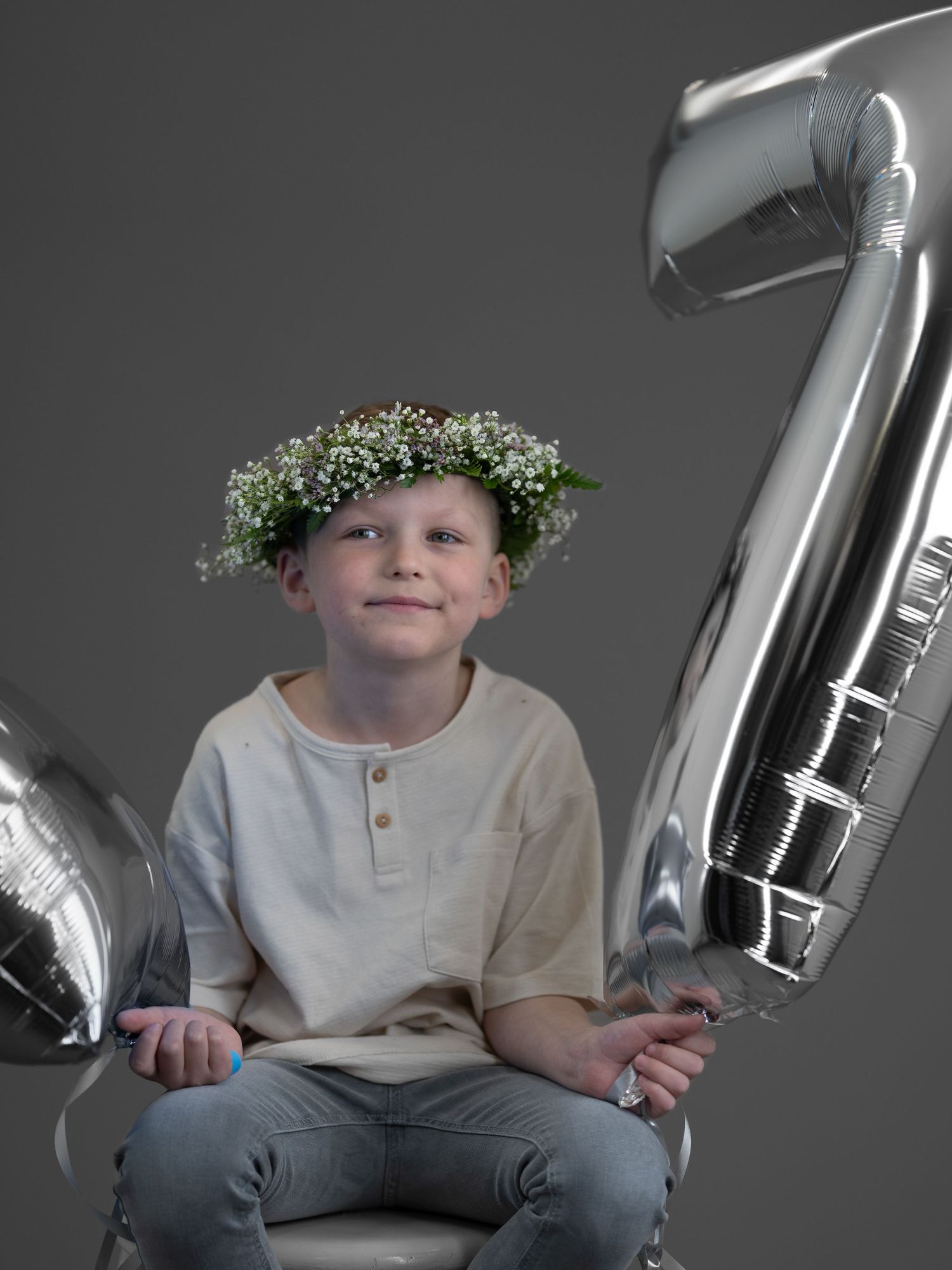 A child wearing a white flower crown and light colored shirt sits with silver number balloons against a gray background.