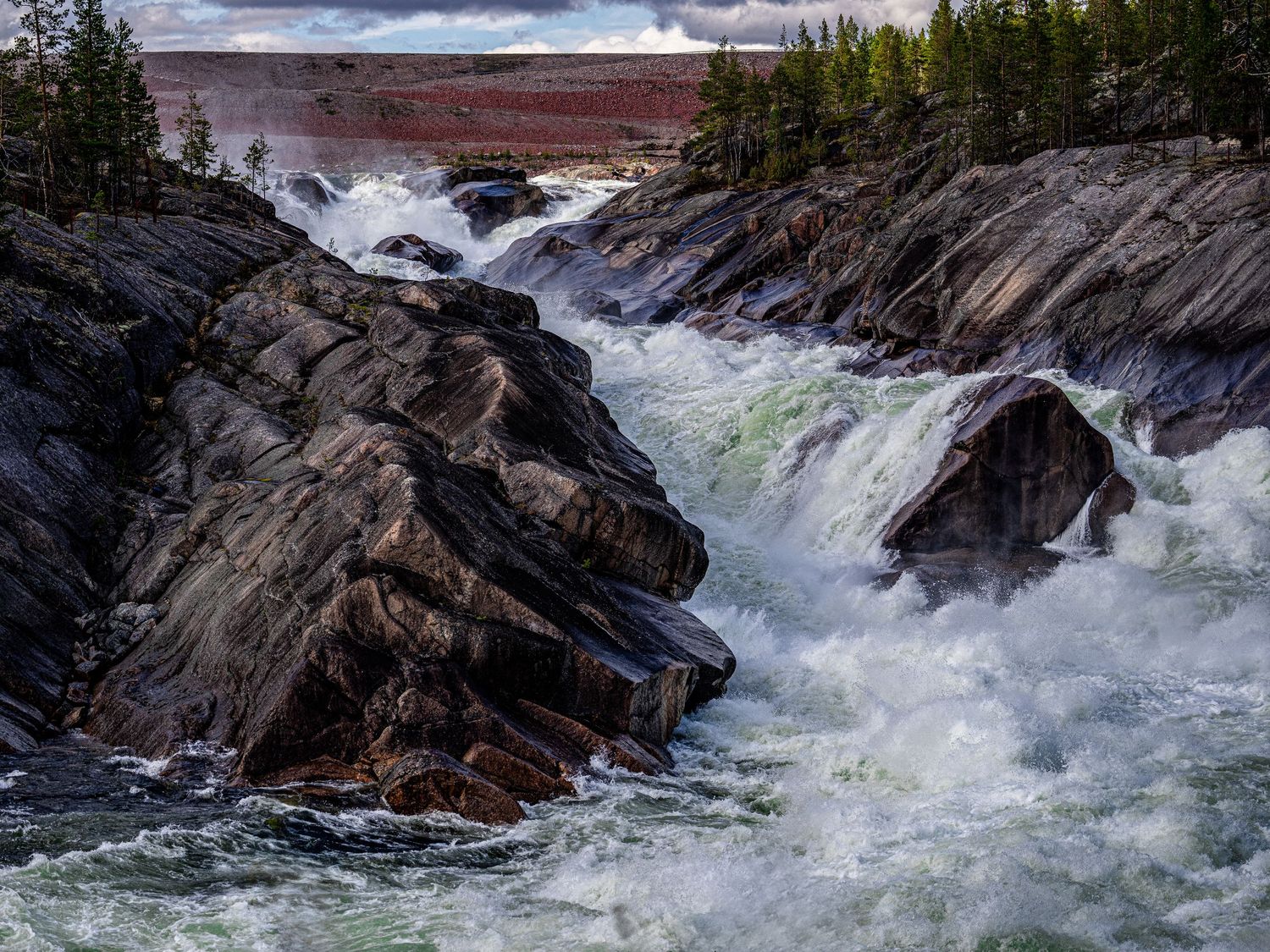 Rushing whitewater rapids flow between jagged rocks in a scenic wilderness river landscape.