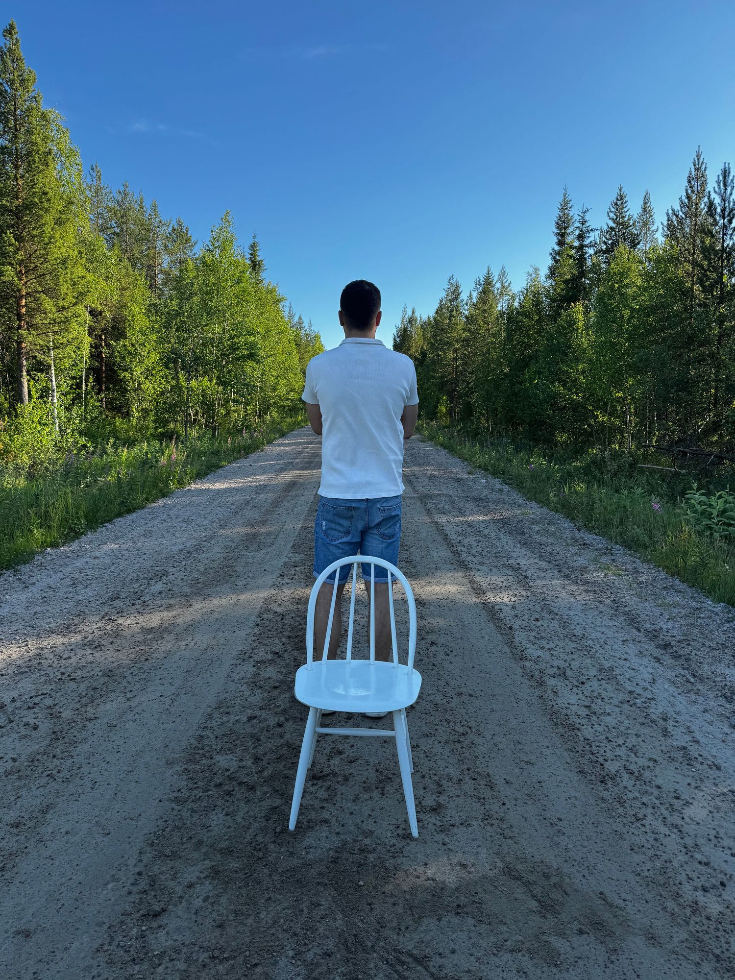 Someone standing on a gravel road with an empty white chair, surrounded by green trees on a sunny day.