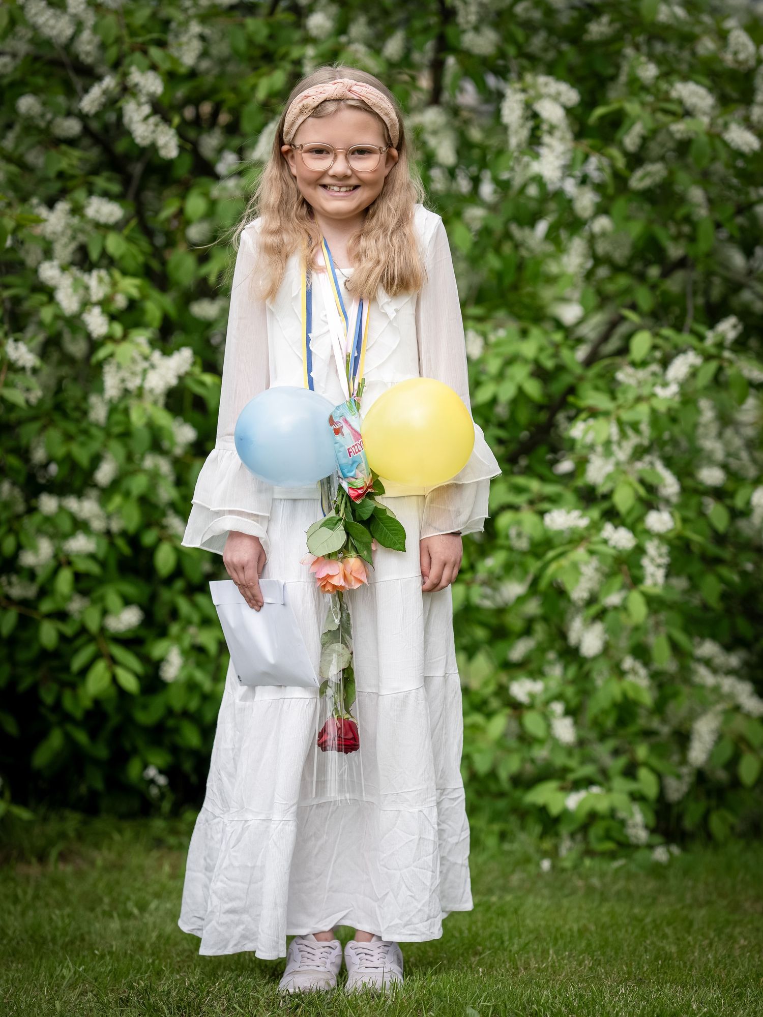 Figur i vit klänning som håller färgglada ballonger framför en blommande vårträdgård.
