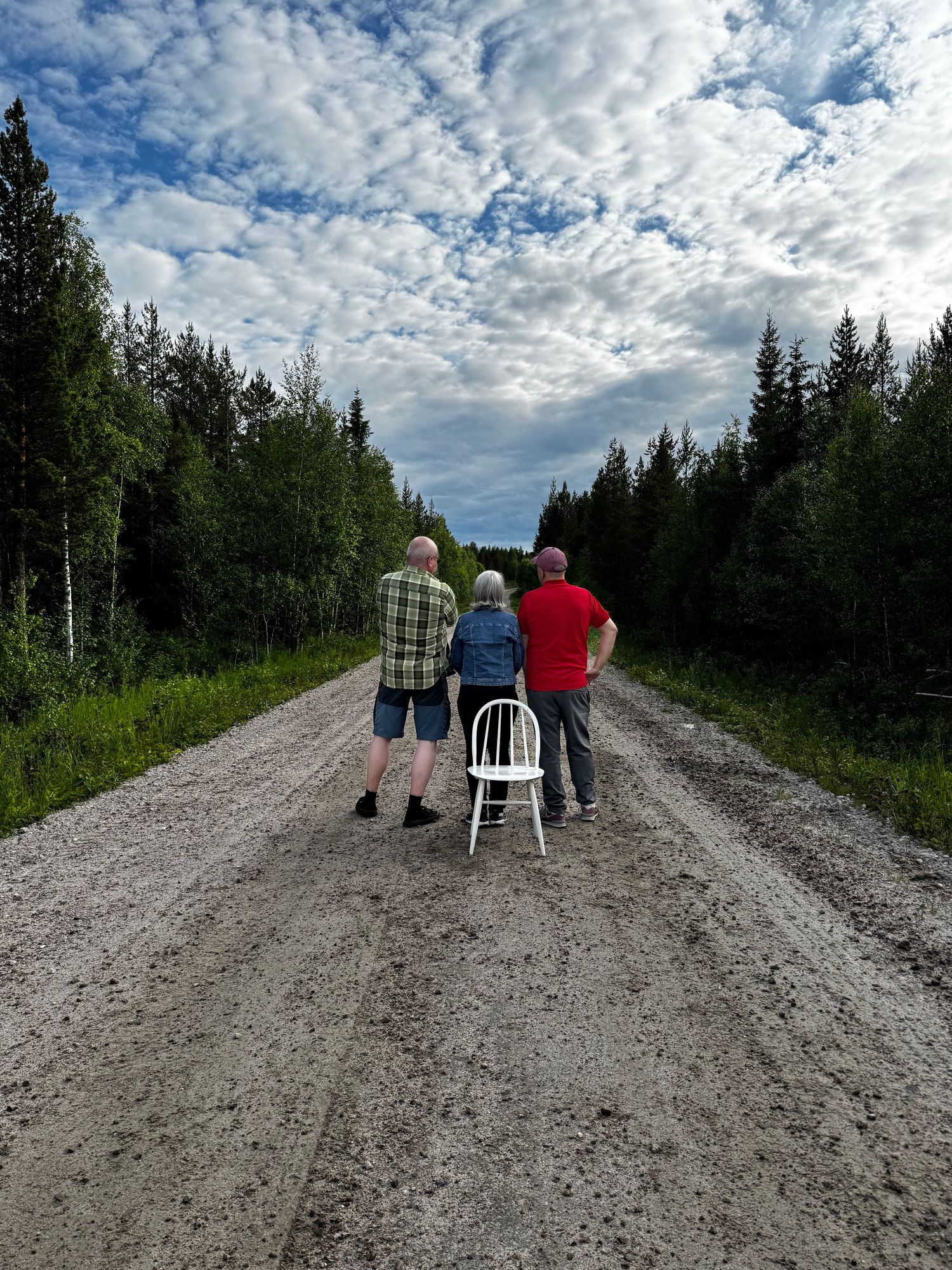 People walking along a rural dirt road bordered by evergreen trees under a dramatic cloudy summer sky.