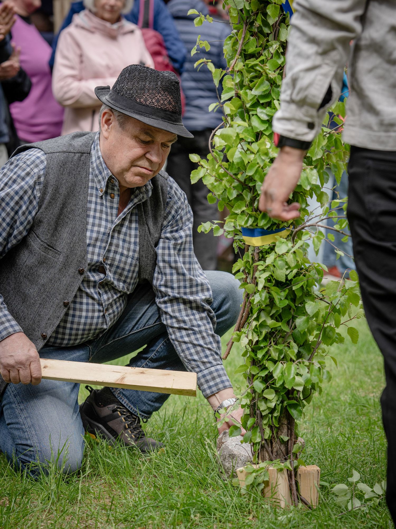 Person wearing a hat and checkered shirt kneels by greenery during an outdoor garden demonstration.