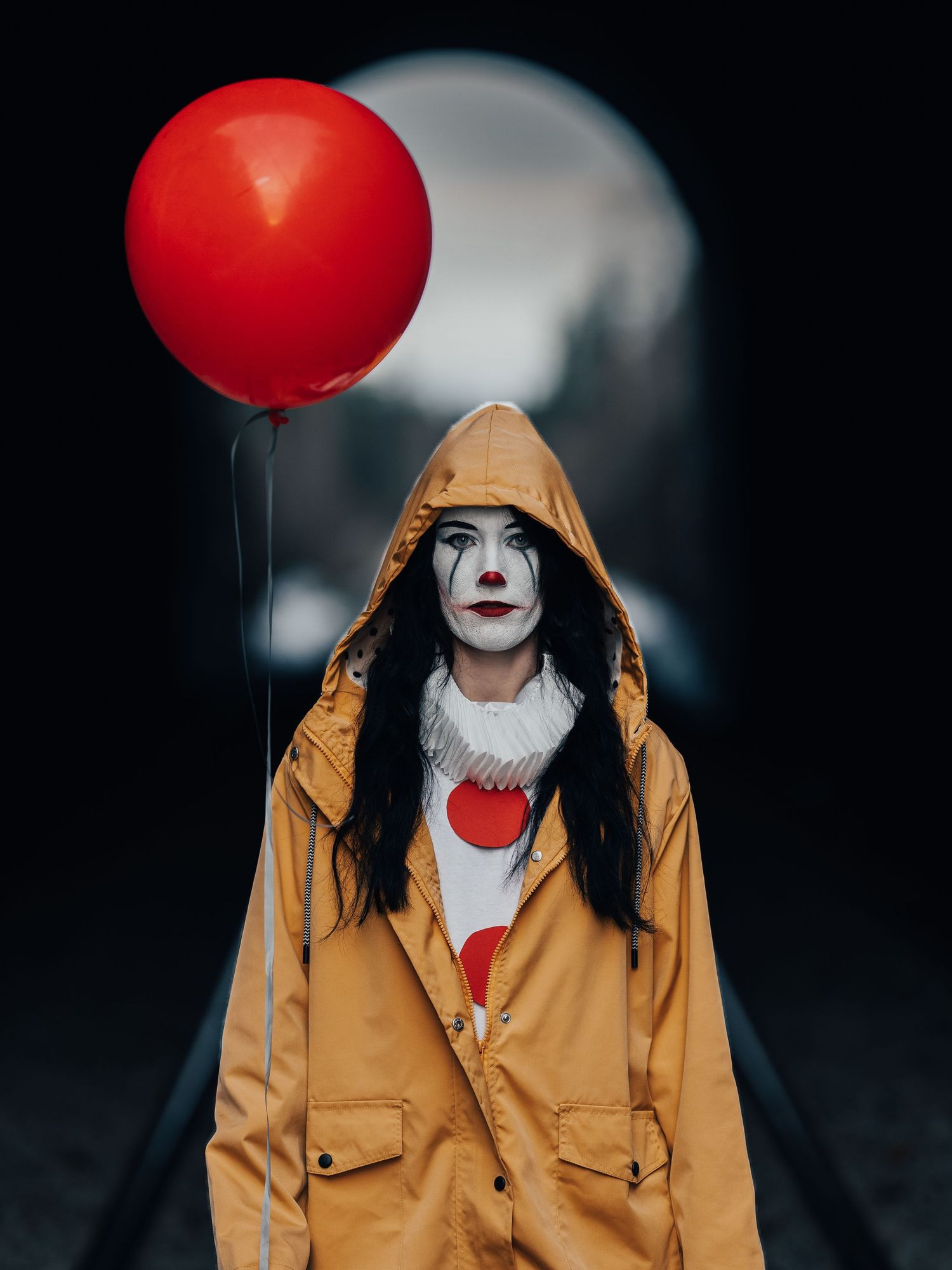 Mörkt konstnärligt porträtt av clownfigur i gul rock med röd ballong mot skuggig bakgrund.