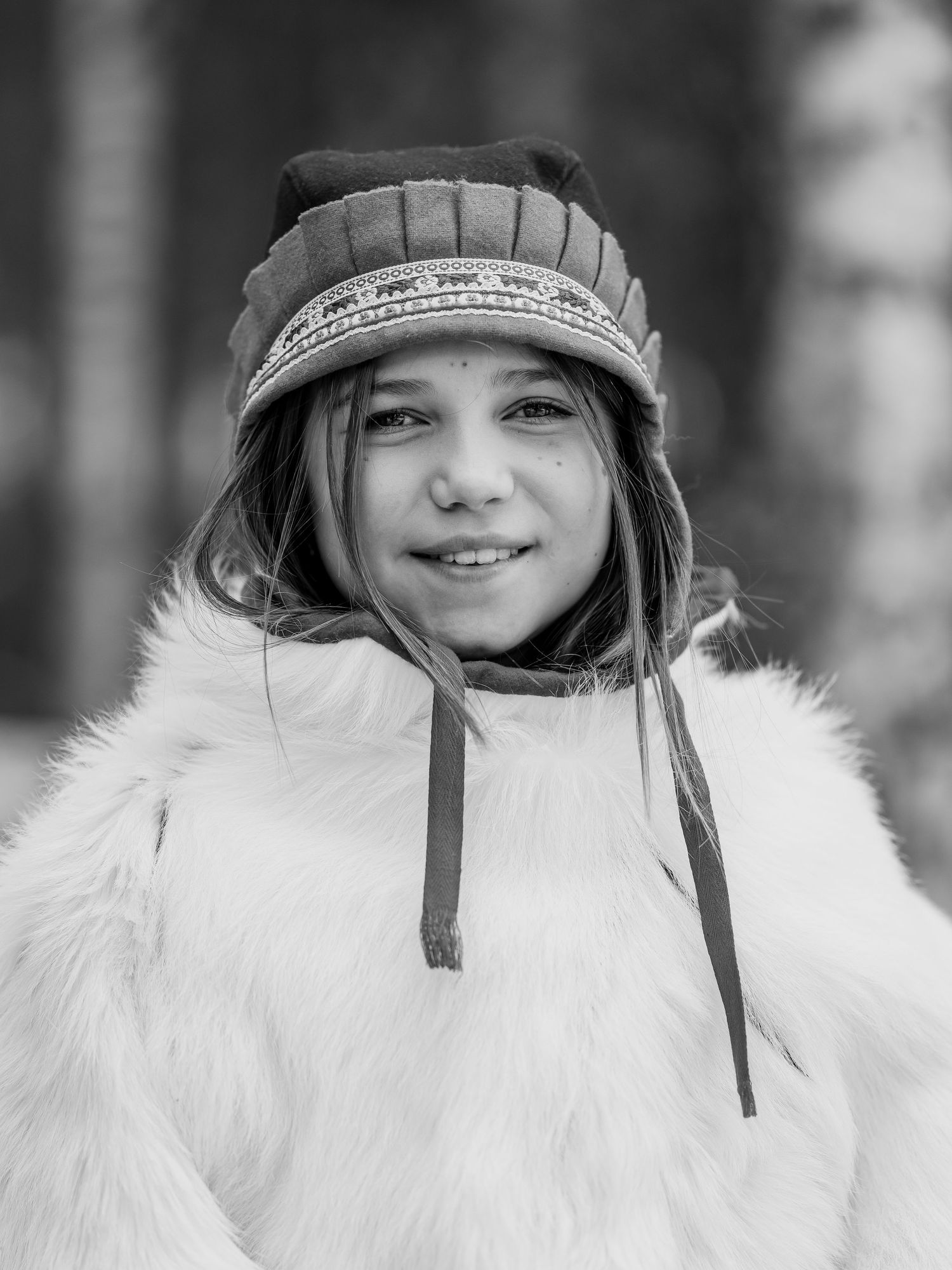 Vinterporträtt av person i vit pälskappa och mönstrad hatt. Samisk klädsel.