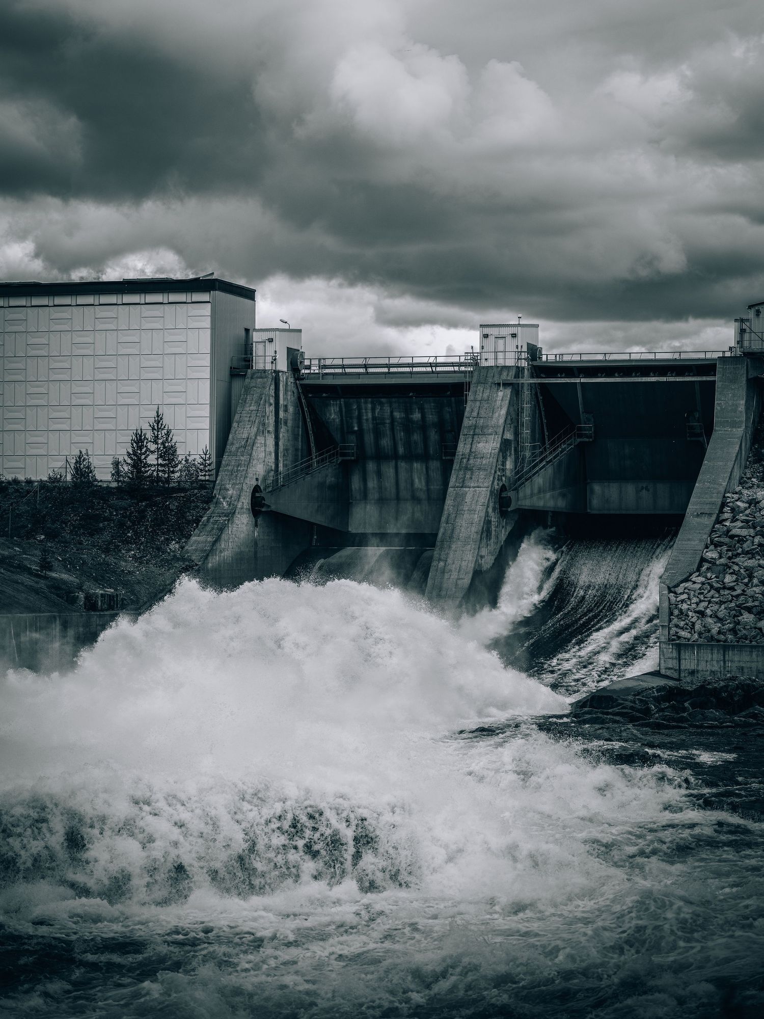 A dramatic hydroelectric dam with water rushing through its spillways under dark storm clouds, captured in monochrome.