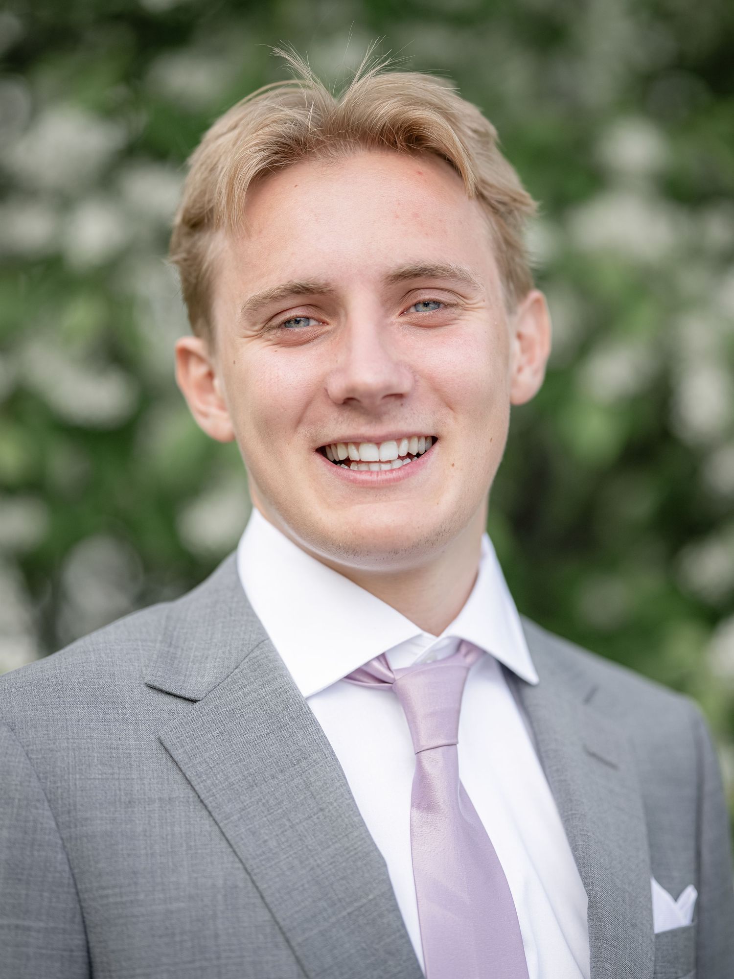 Formal portrait wearing a gray suit with lavender tie against a blurred floral background.