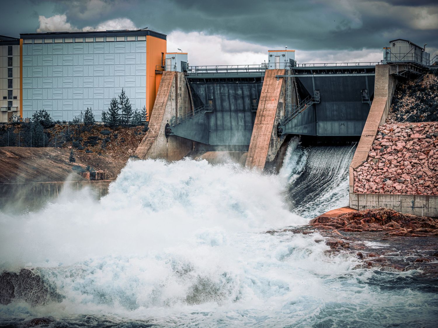 Powerful hydroelectric dam releases turbulent white water through its gates under a dramatic stormy sky.