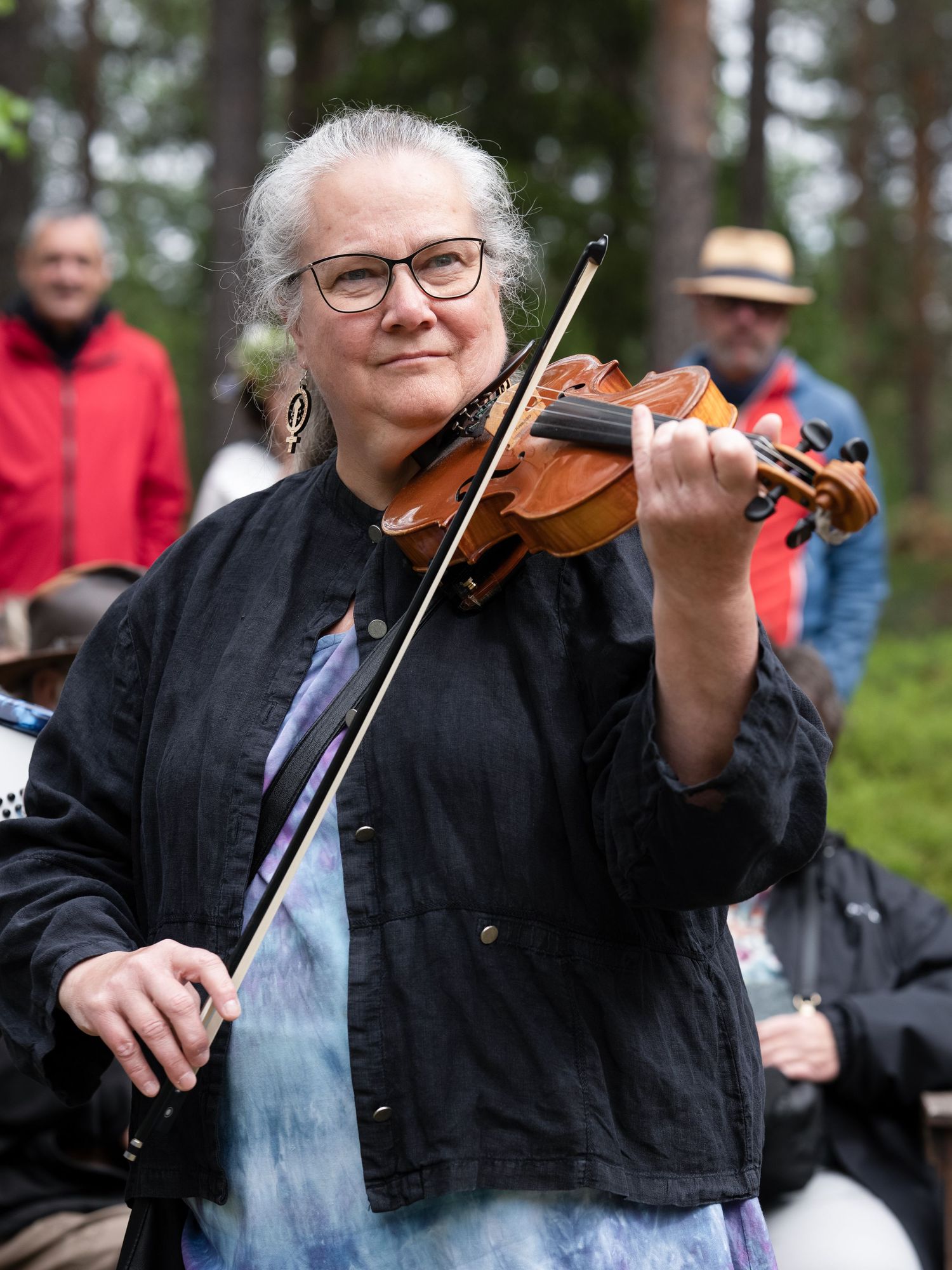 A violinist performs outdoors wearing a black jacket and glasses, with people visible in the background.
