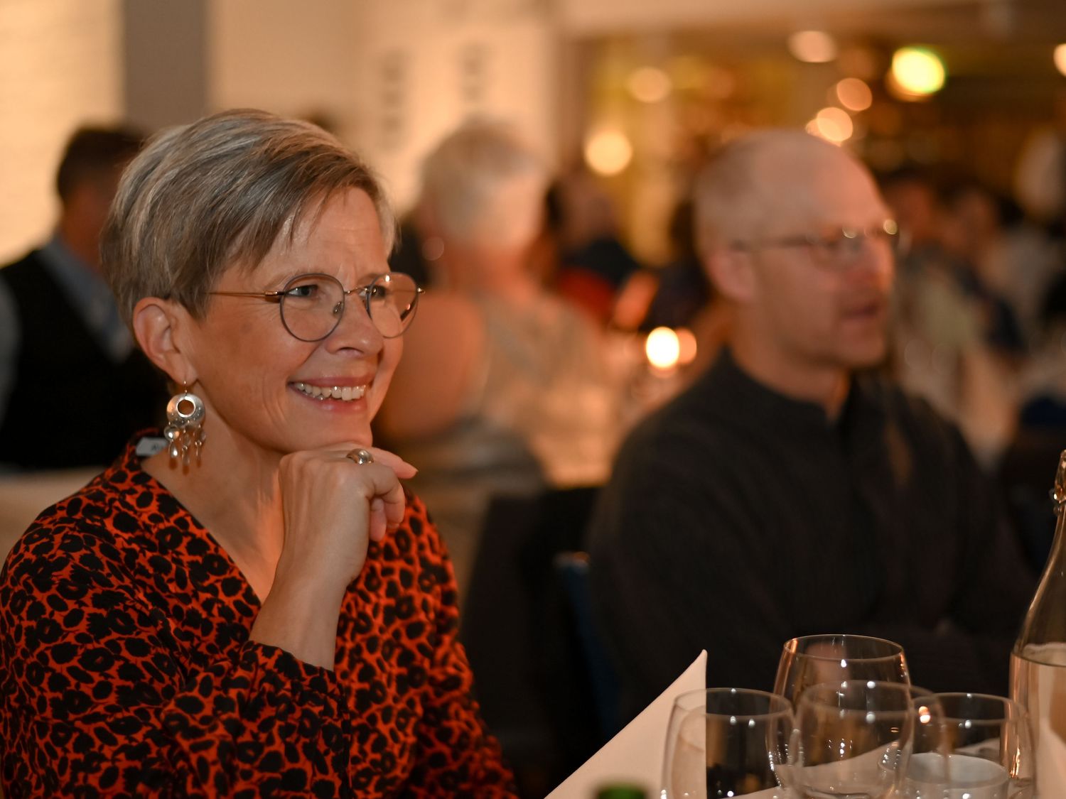 Someone in glasses and a red patterned top smiles while dining at a restaurant with warm ambient lighting in the background.