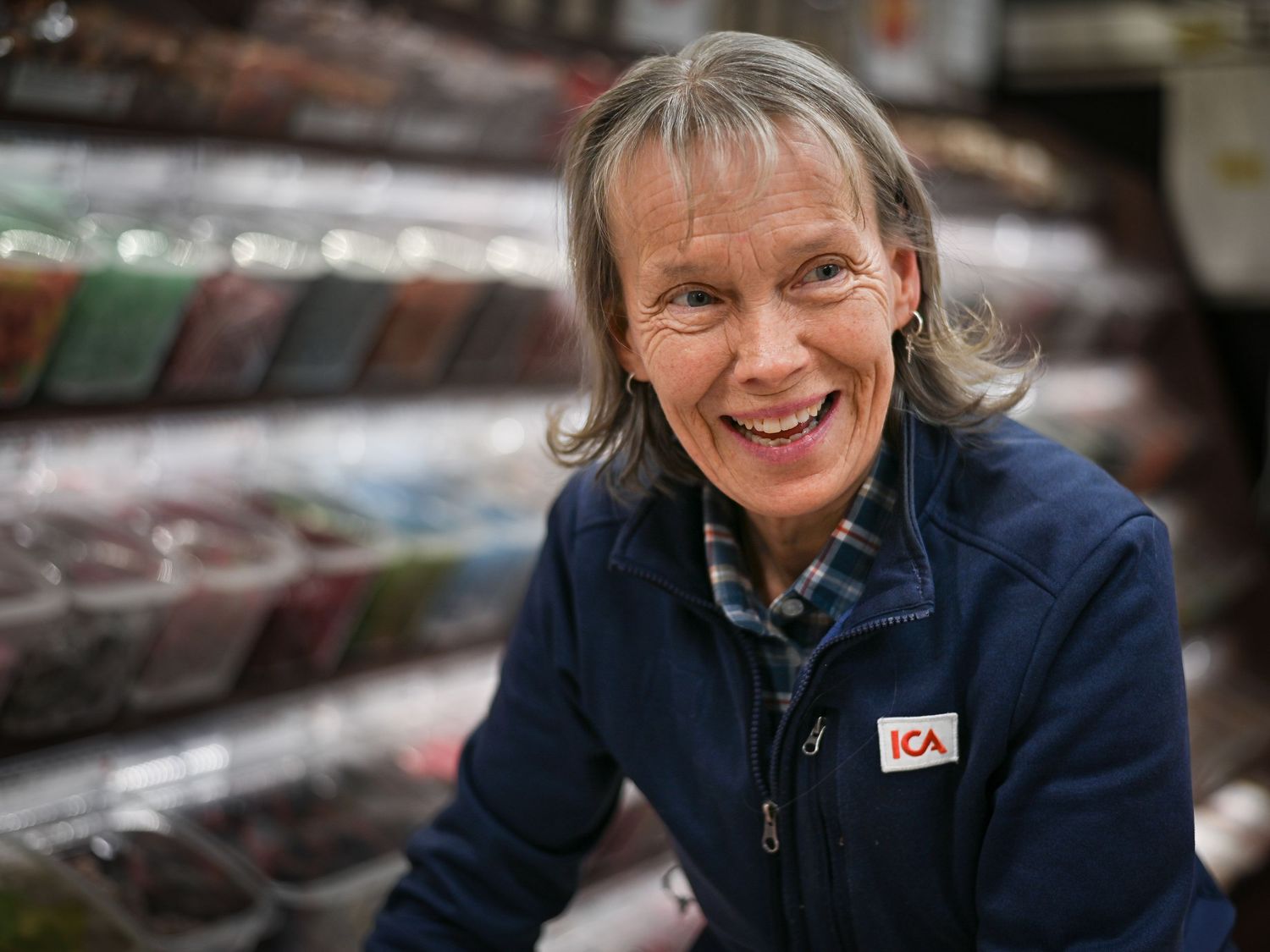 ICA-butiksanställd i marinblå jacka ler varmt medan de arbetar i butiksmiljö.