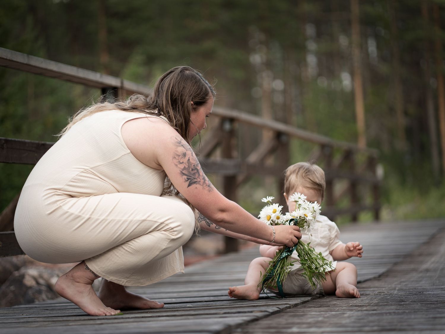 Person i ljus klänning hukar sig bredvid ett litet barn med blommor på en trägångväg.