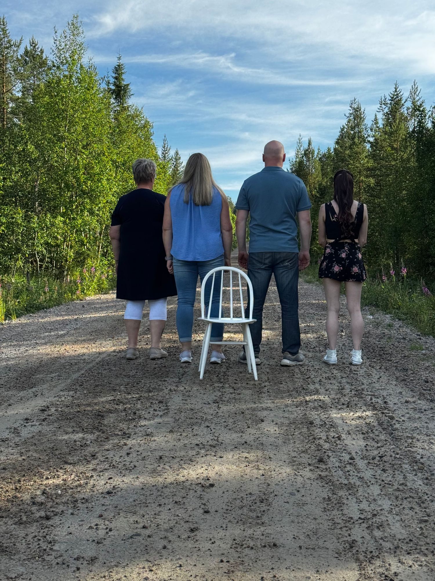 Four people stand on a dirt path facing a scenic forest view with a single white chair placed behind them.
