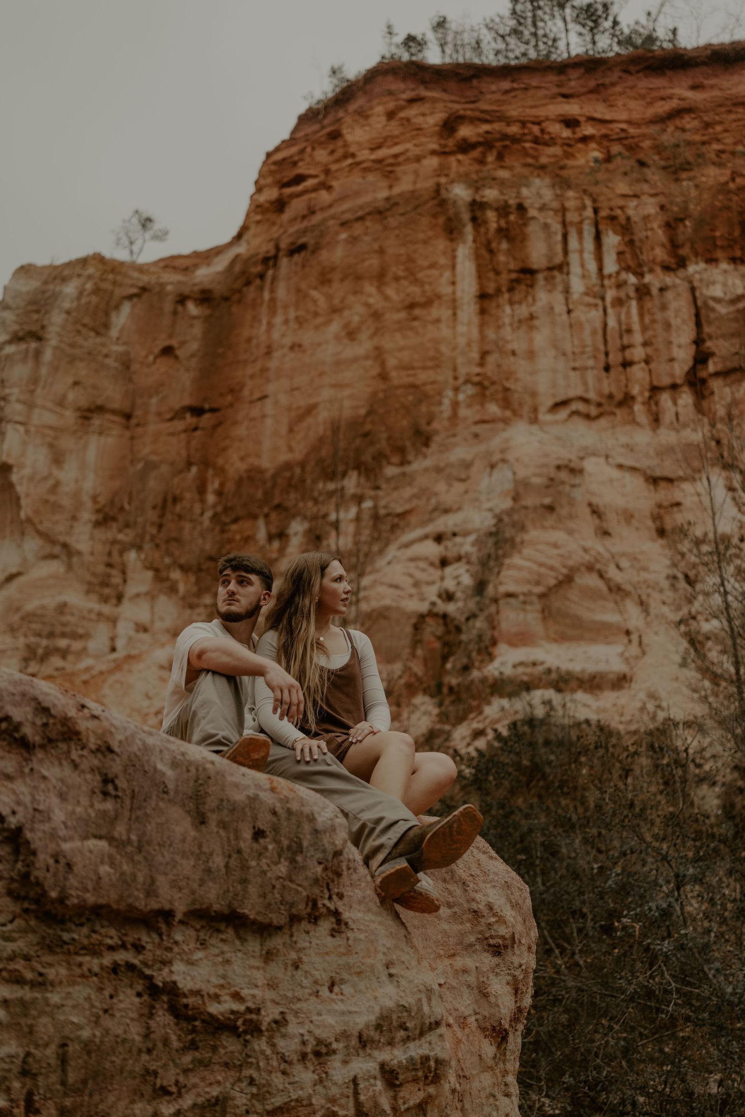 Couple sitting on rocky cliff edge overlooking natural canyon landscape at sunset.