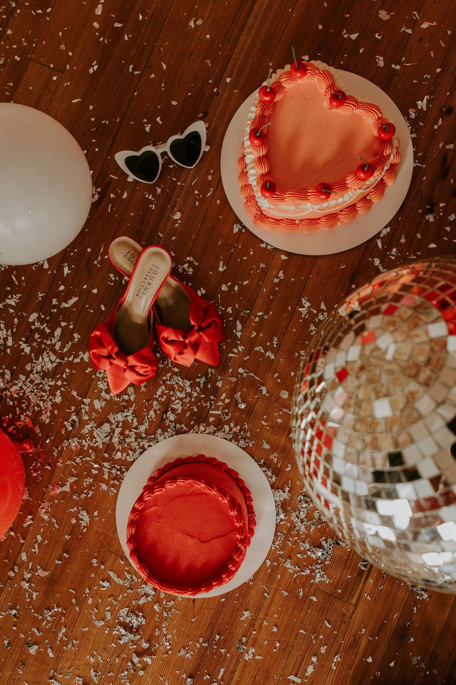 Heart-shaped red cakes, heart-shaped sunglasses, and a disco ball on wooden surface with confetti scattered around.
