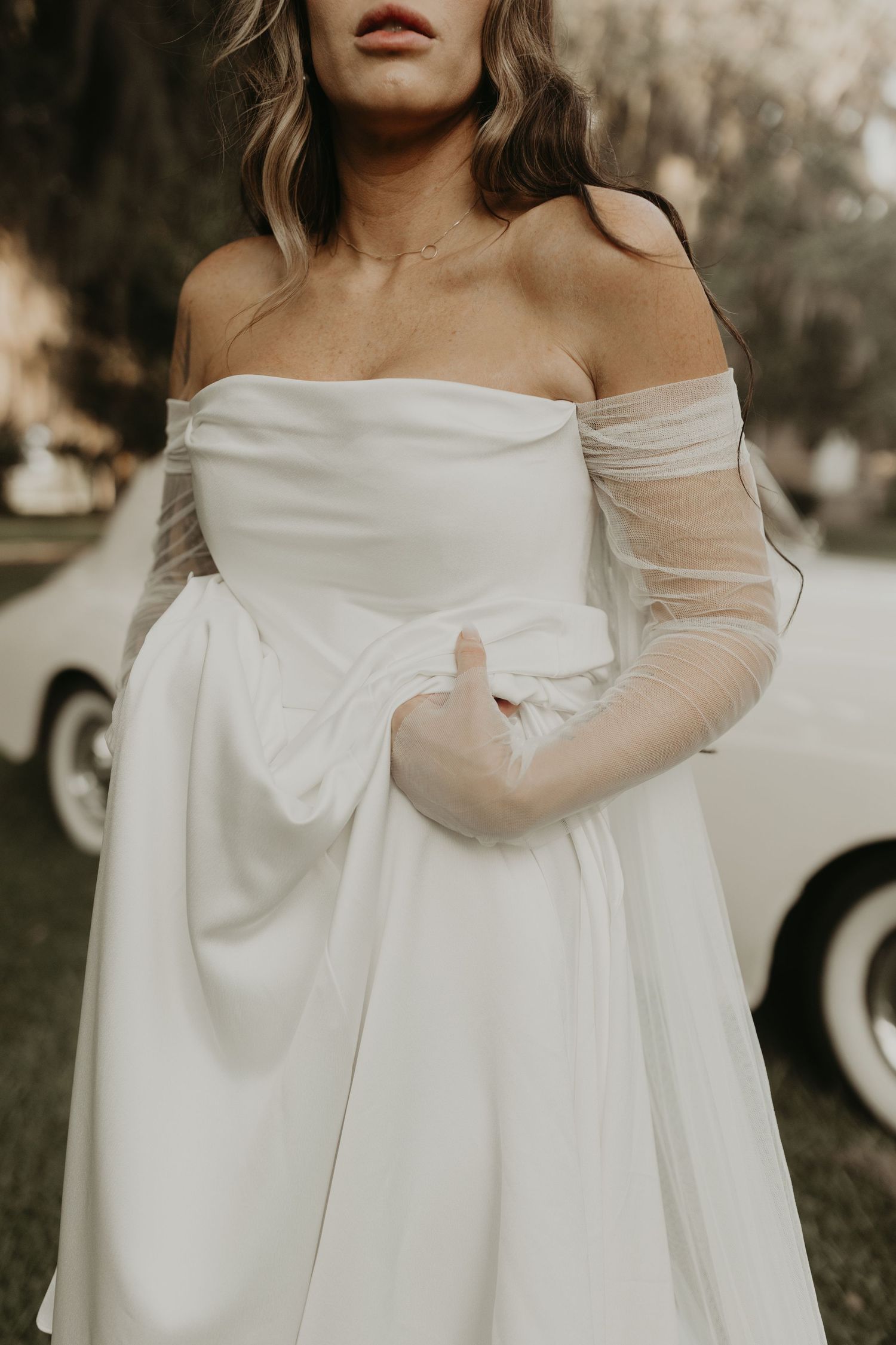 Detail shot of off-shoulder white wedding dress with sheer sleeves against vintage setting.