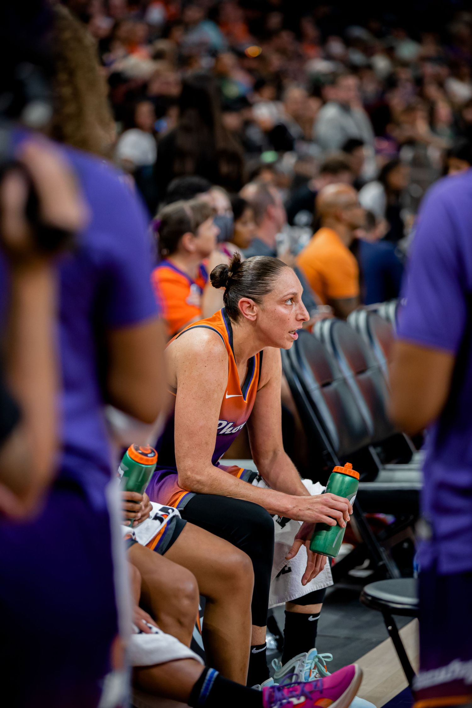 Basketball player in purple uniform sits on sideline bench during game holding water bottles.
