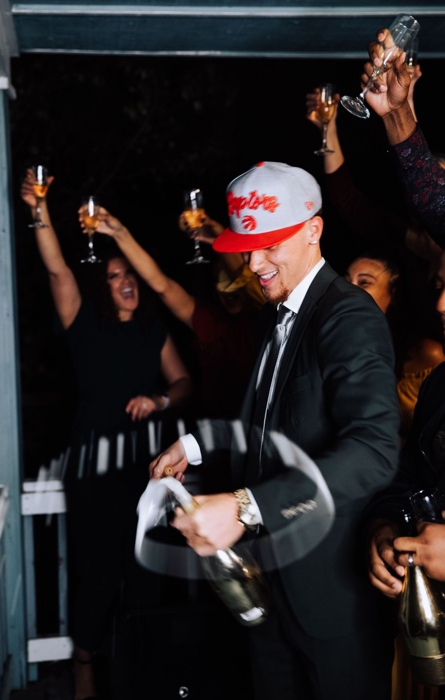Person in suit and baseball cap celebrates with champagne bottles in dark venue.