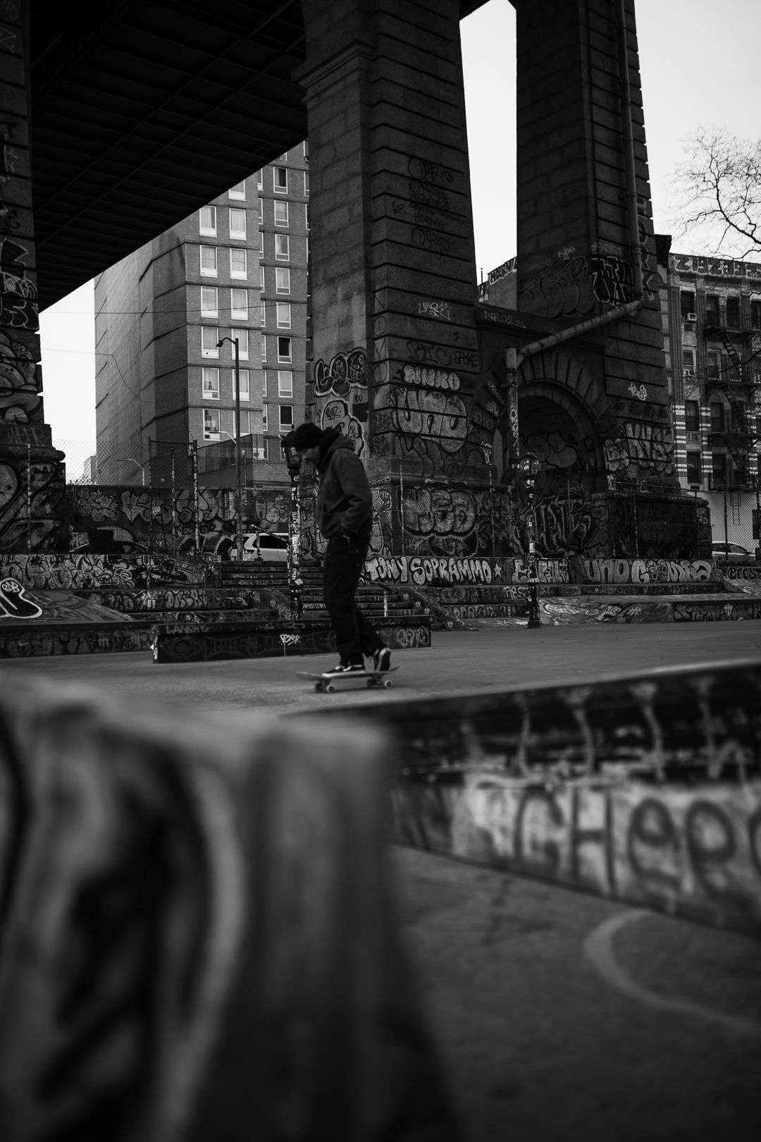 Black and white urban skateboarding scene under bridge with city buildings in background.