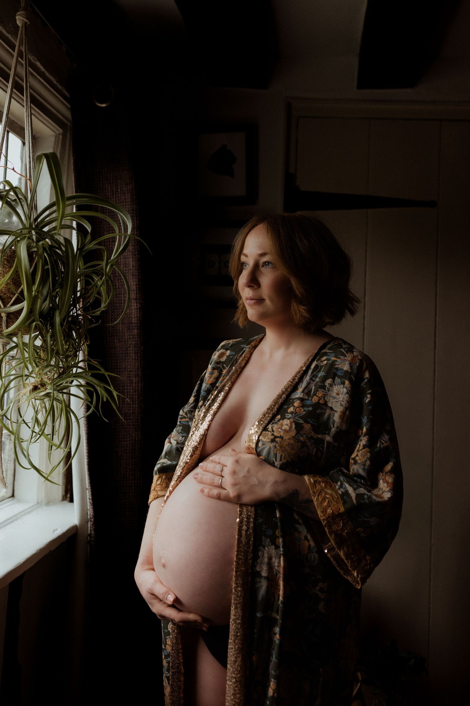 Intimate maternity portrait in dark moody lighting shows expectant figure in patterned robe by window with houseplant.