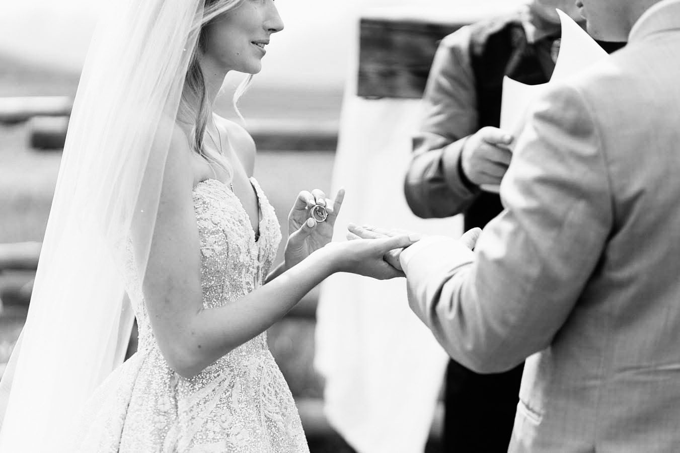 Black and white wedding photograph showing exchange of rings during an outdoor ceremony.