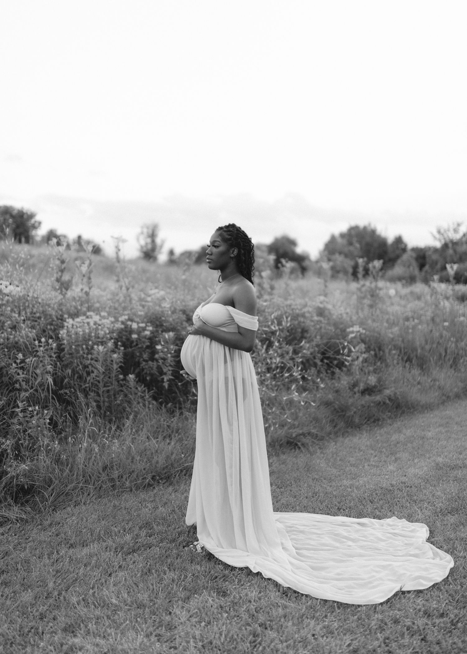 A pregnant woman in a flowing white off-shoulder maternity gown poses in a wildflower field during a black and white photo shoot.