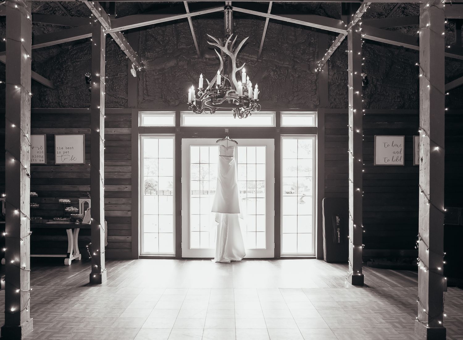 Wedding dress hanging in rustic barn venue with chandelier and string lights.