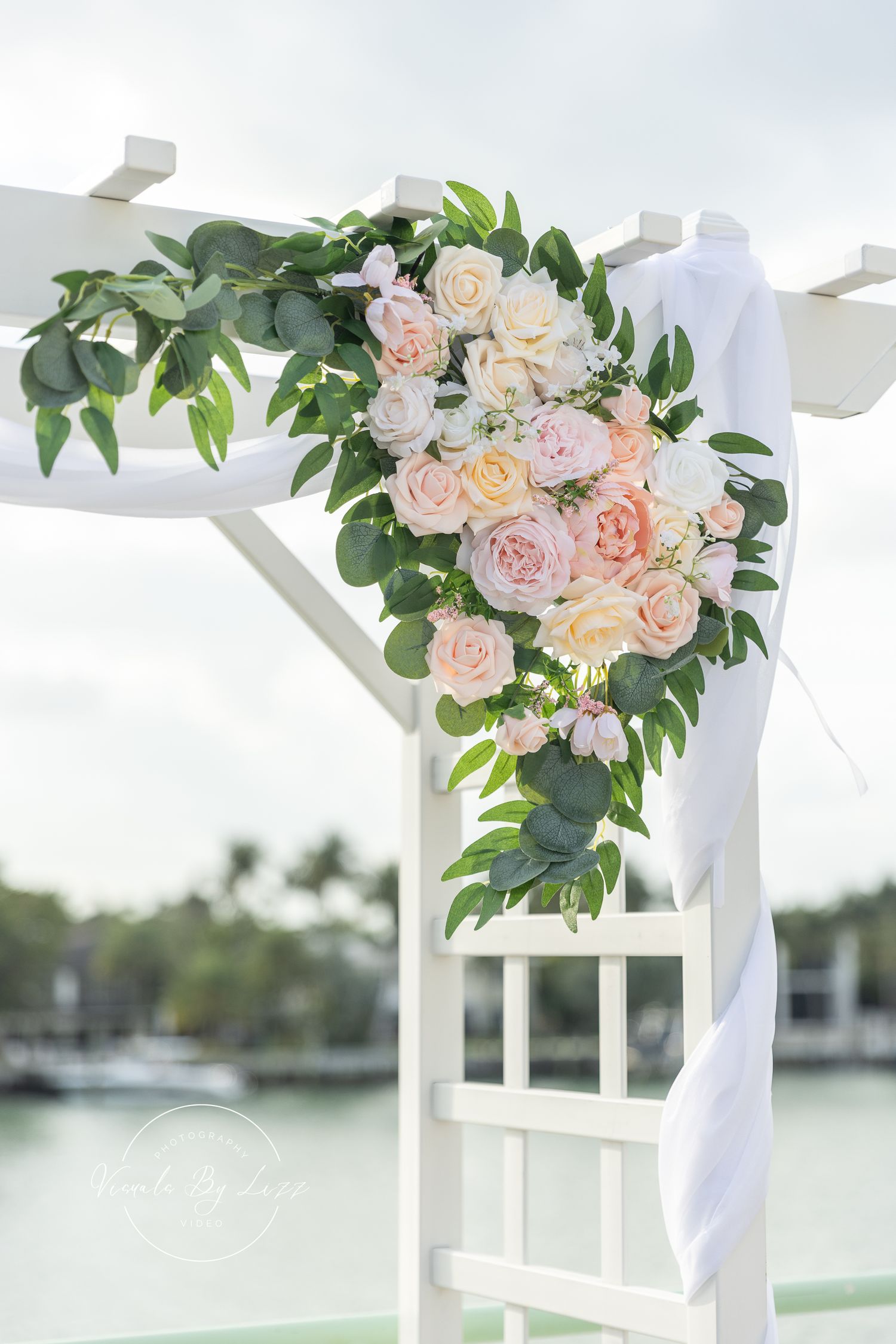 White wedding arch adorned with blush pink roses and greenery against a waterfront backdrop.
