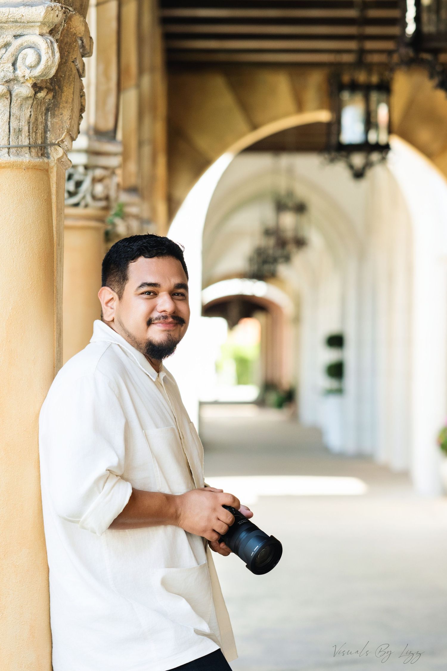 A photographer with a camera stands in an ornate arched hallway with classical stone columns and hanging lanterns.