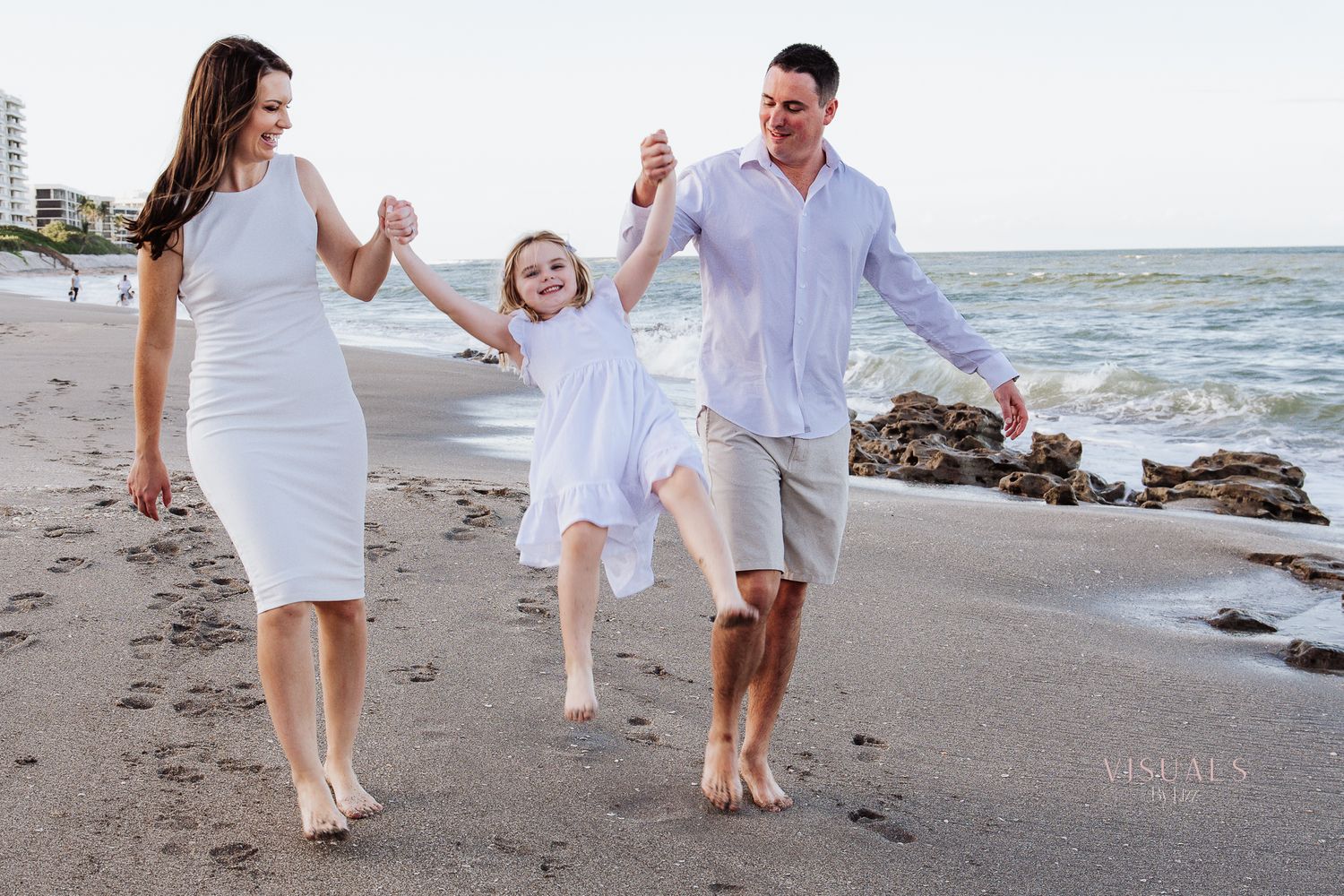 Family in white outfits walking together on beach near ocean during sunset at golden hour.