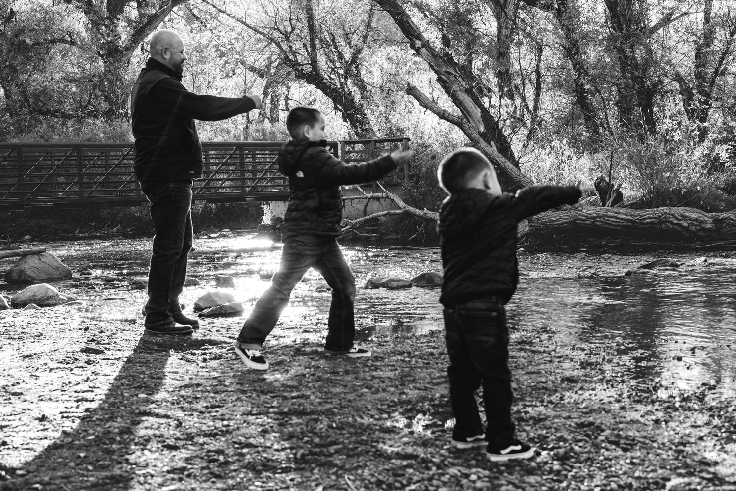 Father and sons playing near river, all three throwing rocks in river.