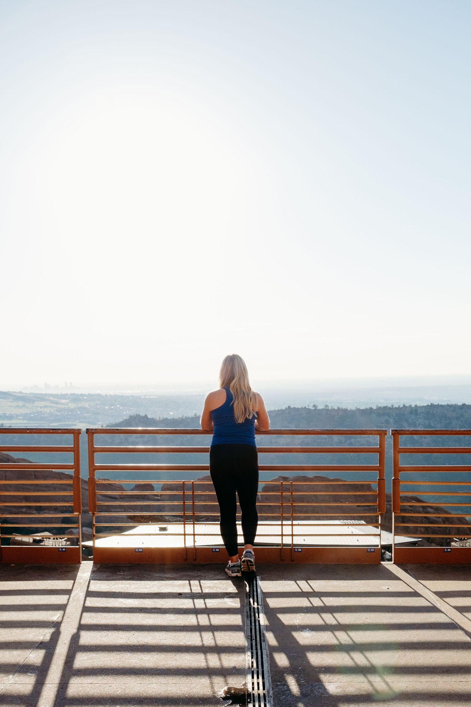 Female trainer standing against railing at Red Rock Amphitheatre, sun shining brightly with slight Denver city scape views in background.