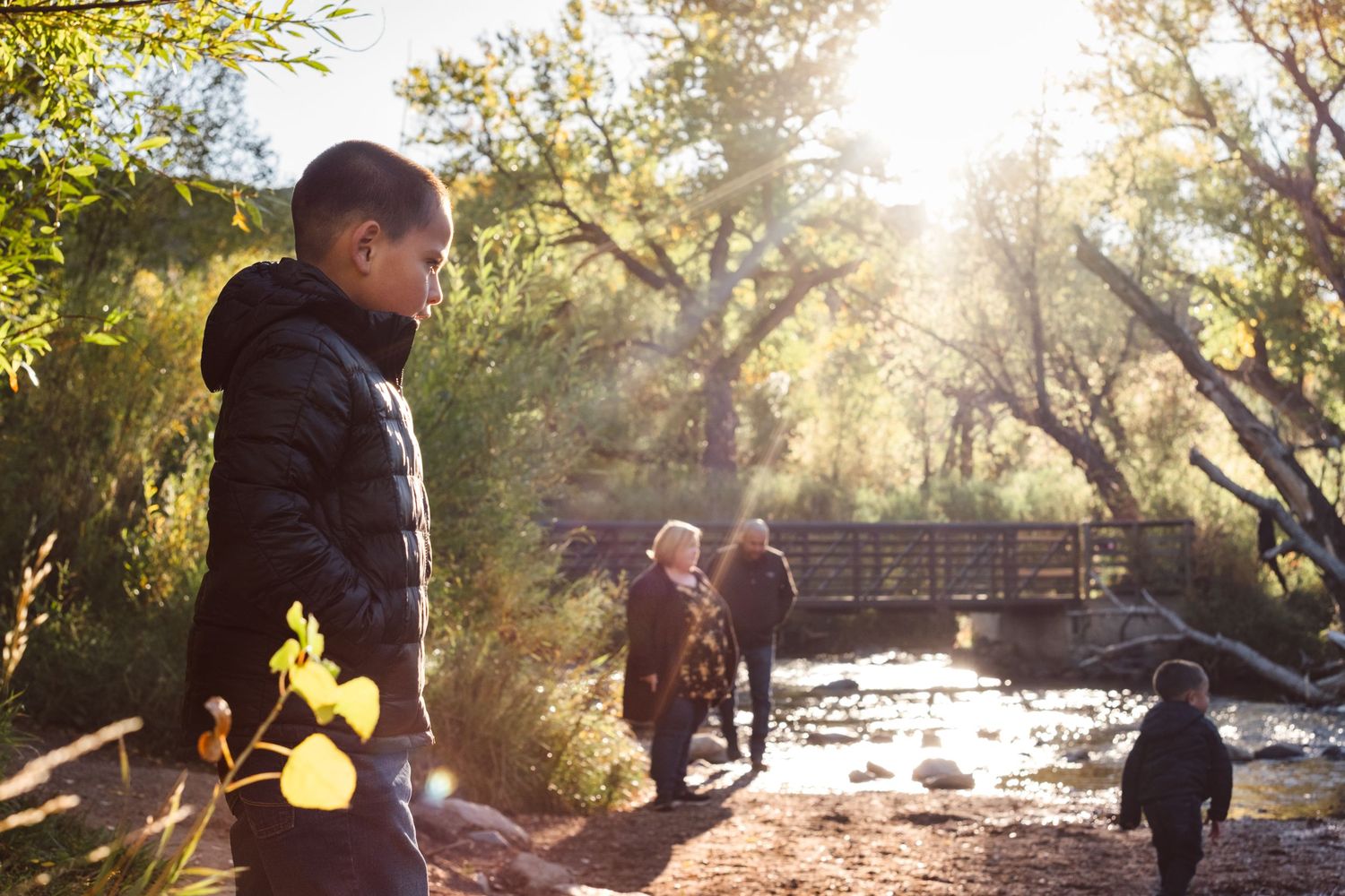 Documentary photo of family playing near river, bridge in background, sun beaming through fall trees.