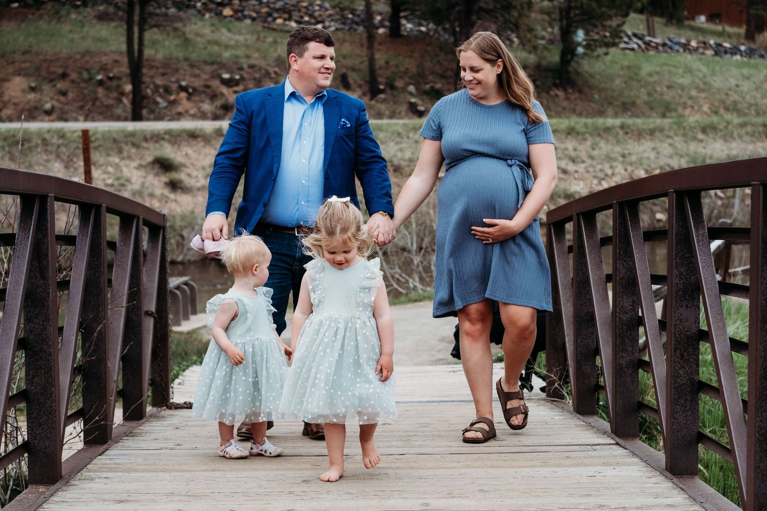 Heartfelt photo of father looking toward pregnant mother, mother smiling down to toddler girls on boardwalk bridge in Evergreen.