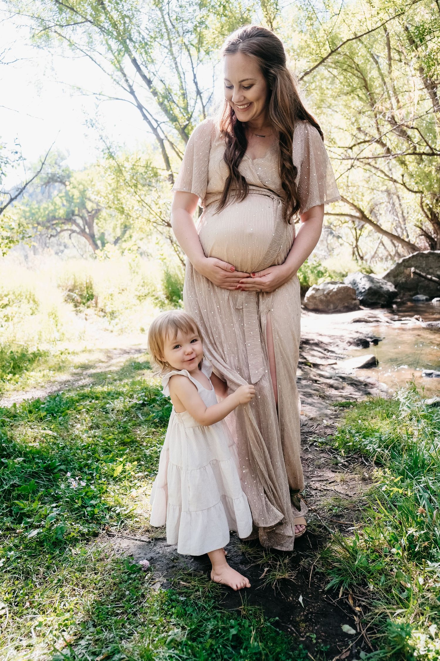 Pregnant mother wearing champagne colored dress, toddler daughter hugging her leg, laughing.