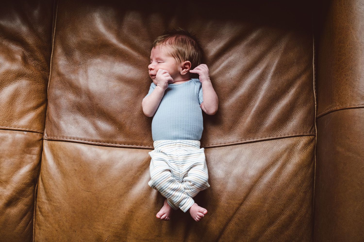 Newborn baby boy scrunched up on camel color couch.