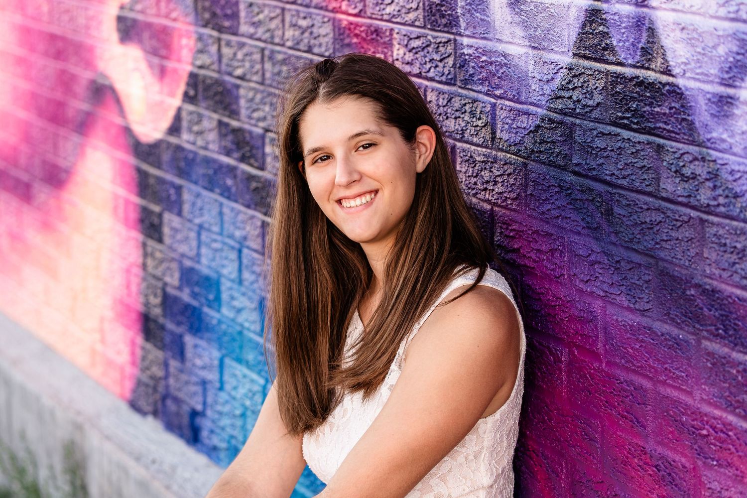 Teenage girl wearing white dress, smiling next to colorful mural painting on brick building.