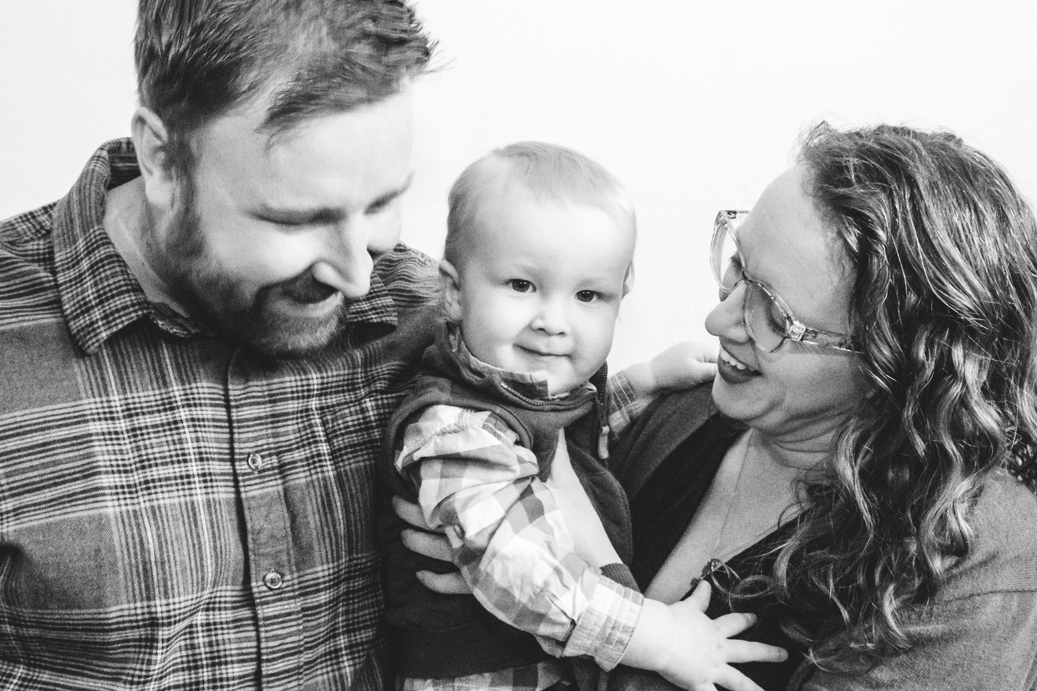 Studio portrait of mother holding toddler son, standing close to father, both mother and father smiling at toddler.