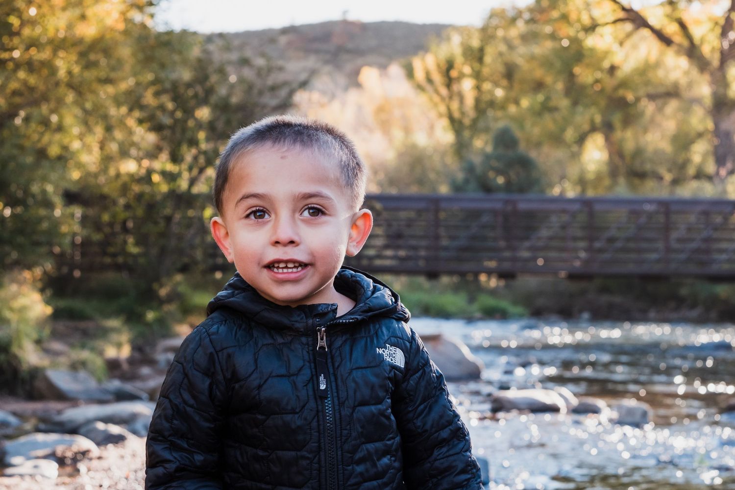Playful portrait of young boy standing in front of river at Lair o the Bear, bridge in background.