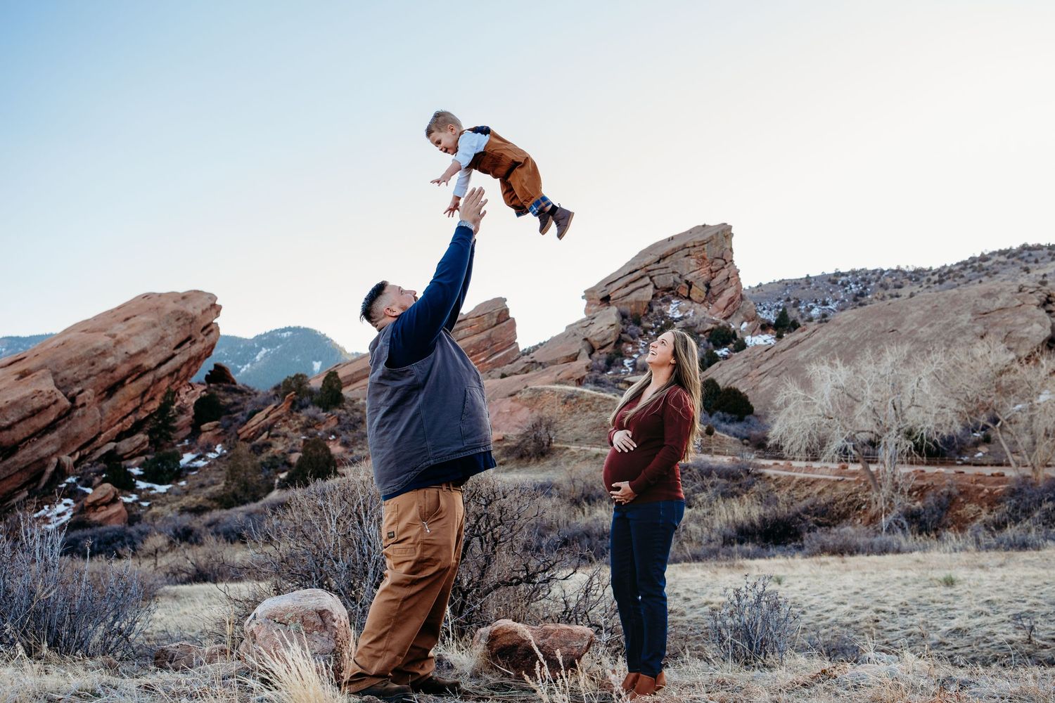 Father tossing toddler into air, pregnant mother standing nearby watching in Red Rocks Park, Morrison Colorado.